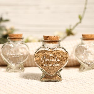 Pode incluir: Três frascos de vidro em forma de coração com tampas de cortiça. Os frascos estão cheios de areia e têm texto personalizado na frente. O texto nos frascos diz "Emily & John 16.08.25", "The Amanda 02.12.2025" e um desenho floral com a data "16.08.25".