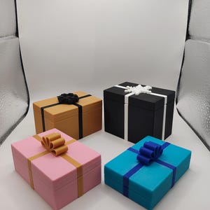 May include: Four rectangular gift boxes in various colors: tan, black, pink, and teal. Each box is adorned with a contrasting ribbon and bow. The tan box has a black bow, the black box has a white bow, the pink box has a gold bow, and the teal box has a blue bow.