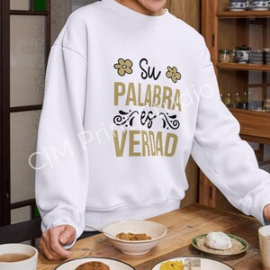 May include: White sweatshirt with the Spanish text "Su Palabra es Verdad" in gold and black lettering. The design includes floral accents. The sweatshirt is displayed on a wooden table.