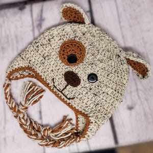 May include: A handmade crochet hat in the shape of a dog's head. The hat is primarily beige with brown accents, including the ears, nose, and a large spot. Black buttons form the eyes. Braided ties with tassels complete the design.
