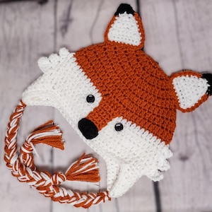 May include: A handmade crochet fox hat in orange, white, and black. The hat features a fox face with button eyes, a black nose, and pointed ears. Braided ties with tassels complete the design. The hat is designed for children.