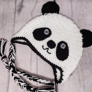 May include: A handmade crochet panda hat in white and black. The hat features black panda eyes, ears, and a nose, with a stitched mouth. Braided black and white ties hang from the hat. The hat is designed for children.