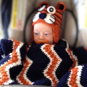 May include: A baby wearing a crocheted orange and black tiger hat with matching blanket. The blanket has a chevron pattern in navy blue, orange, and white. The baby is in a car seat.