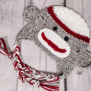 May include: A handmade crochet sock monkey hat in gray, white, and red. The hat features a white face with a red mouth, black button eyes, and a white and red striped top. A braided tassel hangs from the hat.