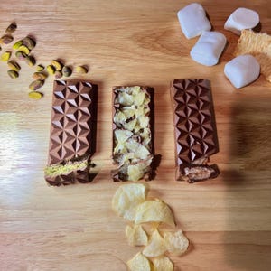 Peut inclure: Trois barres de chocolat avec des motifs géométriques, chacune partiellement cassée pour révéler différentes garnitures : pistaches, chips de pommes de terre et une garniture crémeuse. Pistaches, chips de pommes de terre, guimauves et craquelins éparpillés.