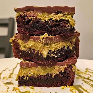May include: Three stacked brownies with layers of chocolate, a light green filling, and a chocolate topping. The brownies are on a white plate with green drizzle. The dessert is a close-up shot.