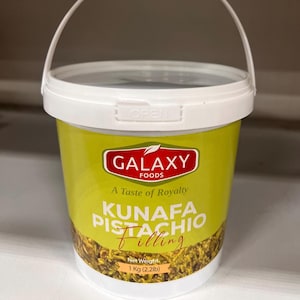May include: A white plastic bucket with a yellow label that says "Galaxy Foods, A Taste of Royalty, Kunafa Pistachio Filling, Net Weight 1 Kg (2.2lb)". The bucket is filled with pistachio filling.