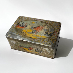 May include: Vintage metal tin box featuring a colourful illustration of people in a garden setting. The box has a worn, aged look with a faded yellow, red, and green colour scheme.