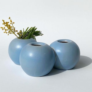 May include: Three light blue ceramic vases in a round shape. The vases are arranged in a row, with the middle vase slightly higher than the other two. The vases are smooth and have a glossy finish.