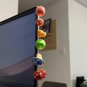 May include: A series of colorful, mushroom-shaped magnets attached to the side of a laptop. The magnets are various colors, including red, orange, green, and blue, with white spots. The mushrooms are a whimsical and decorative item.