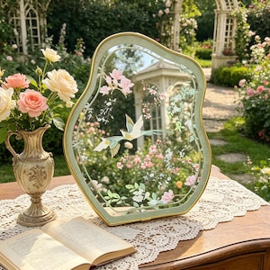 May include: An irregular, organic-shaped tabletop mirror with a sage green frame and gold trim. The glass surface features a decorative print of two flying birds surrounded by delicate pink blossoms and green foliage, reflecting a garden scene.