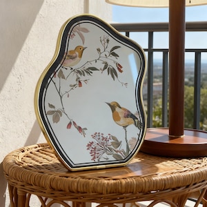 May include: A decorative mirror with a distinctive, wavy outline. It has a gold frame, a black border, and a white interior with a floral and bird pattern. The mirror is displayed on a woven side table.