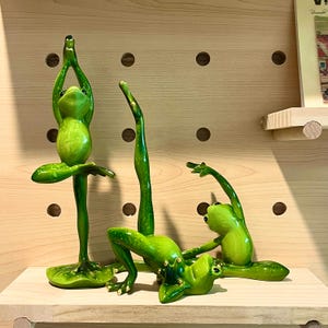 Könnte beinhalten: Drei grüne Froschfiguren in verschiedenen Yoga-Posen. Die Frösche sind aus Keramik und haben realistische Details. Die Figuren werden auf einem Holzregal ausgestellt.