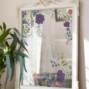 Puede incluir: Un espejo con marco blanco con calcomanías florales. La superficie del espejo está decorada con ilustraciones botánicas coloridas, que incluyen flores moradas, rosas y azules, y follaje verde. El marco tiene detalles ornamentados.