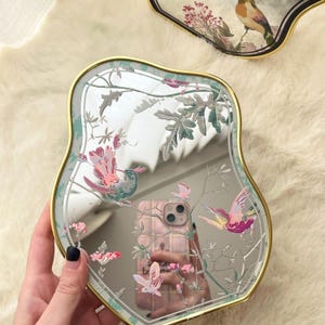 Może przedstawiać: Ozdobne lustro ze złotą obwódką i unikalnym kształtem gruszki. Lustro posiada kolorowy wzór ptaków i kwiatów w odcieniach różu, zieleni i bieli. Lustro trzymane jest w dłoni.