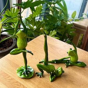 May include: Three green frog figurines in different yoga poses. The frogs are painted with realistic details and have gold accents. The frogs are on a wooden surface.