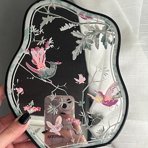 Puede incluir: Un espejo de tocador con marco negro y borde ondulado. El espejo tiene un diseño floral con colibríes rosas y verdes.