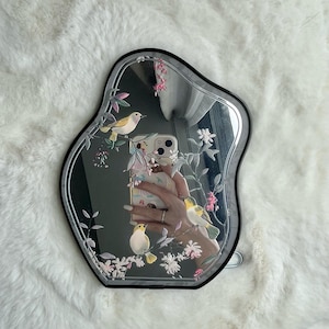 May include: An irregularly shaped mirror with a black frame. The mirror's surface features a delicate design of birds, flowers, and foliage in soft pastel colors. The reflection shows a person holding a phone. The mirror is on a white, fluffy surface.