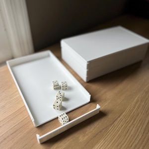 Könnte beinhalten: Ein weißes Würfeltablett mit vier Würfeln und einem Stapel weißer Boxen. Die Würfel sind cremefarben mit schwarzen Punkten. Das Tablett ist rechteckig mit einem erhöhten Rand. Die Gegenstände befinden sich auf einer Holzoberfläche.