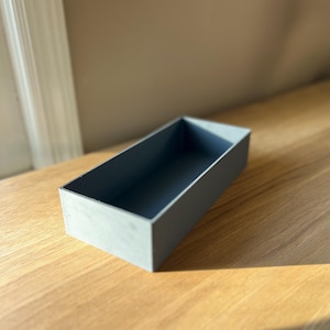 Op de afbeelding: Een rechthoekige, grijze houten doos met een donker interieur. De doos staat op een lichtgekleurd houten oppervlak. De doos is leeg en heeft een eenvoudig, minimalistisch ontwerp.