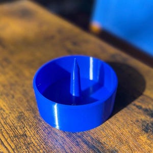May include: A blue ceramic ashtray with a small centre spike. The ashtray is sitting on a wooden surface.