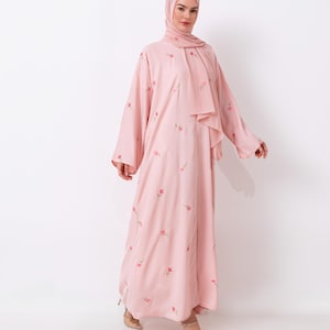 May include: A light pink abaya and hijab set with delicate pink floral embroidery. The long-sleeved abaya has a flowing silhouette, and the matching hijab drapes elegantly. The outfit is paired with nude heels. The logo "Havaa Collections" is in the top left corner.