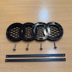 Peut inclure: Emblème Audi en plastique noir avec un motif en nid d'abeille. L'emblème est livré avec quatre vis et deux tiges métalliques noires.