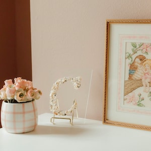 May include: A white ceramic vase with a pink and white checked pattern, filled with a bouquet of pale pink silk flowers. A gold metal stand holds a decorative letter C made of white seashells. A framed cross-stitch embroidery of a bird and a fairy in a pink floral setting.