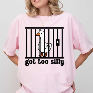 Peut inclure: T-shirt rose clair avec un dessin d'un canard blanc dans une cage noire et l'inscription "got too silly" en noir. La personne porte un short noir. Style d&eacute;contract&eacute;.