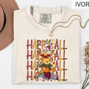 May include: An ivory short-sleeve t-shirt featuring a graphic print of Winnie the Pooh characters including Piglet, Tigger, Pooh, and Eeyore with pumpkins and autumn leaves. The text reads HAPPY FALL in a repeating pattern. The shirt has a Comfort Colors label.