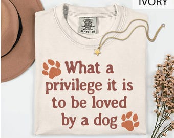 Könnte beinhalten: Ein elfenbeinfarbenes Kurzarm-T-Shirt mit dem Text "What a privilege it is to be loved by a dog" in dunkelorangener Schrift, begleitet von zwei orangen Pfotenabdrücken. Das T-Shirt hat ein Comfort Colors Markenetikett am inneren Halsausschnitt.