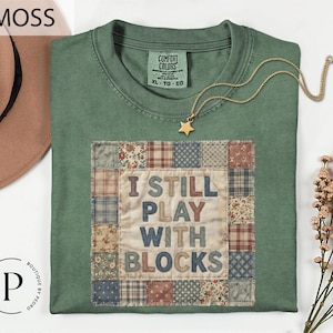 May include: A moss green t-shirt with the text "I STILL PLAY WITH BLOCKS" in a patchwork design. The shirt has a crew neck and short sleeves. A gold star pendant necklace is draped across the shirt. The shirt is size XL-TG-EG.