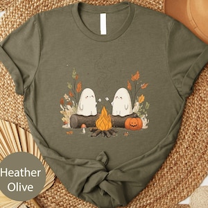 A t-shirt with a graphic design depicting two ghostly figures sitting by a campfire, surrounded by autumn foliage and a pumpkin.