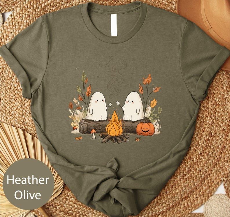 A t-shirt with a graphic design depicting two ghostly figures sitting by a campfire, surrounded by autumn foliage and a pumpkin.