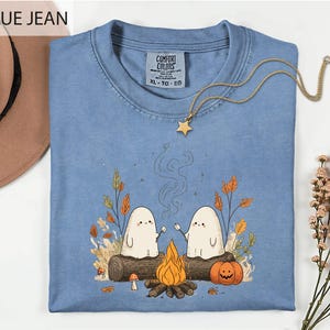 a blue t-shirt with a graphic design depicting two ghostly figures sitting by a campfire, surrounded by autumn foliage and a pumpkin.