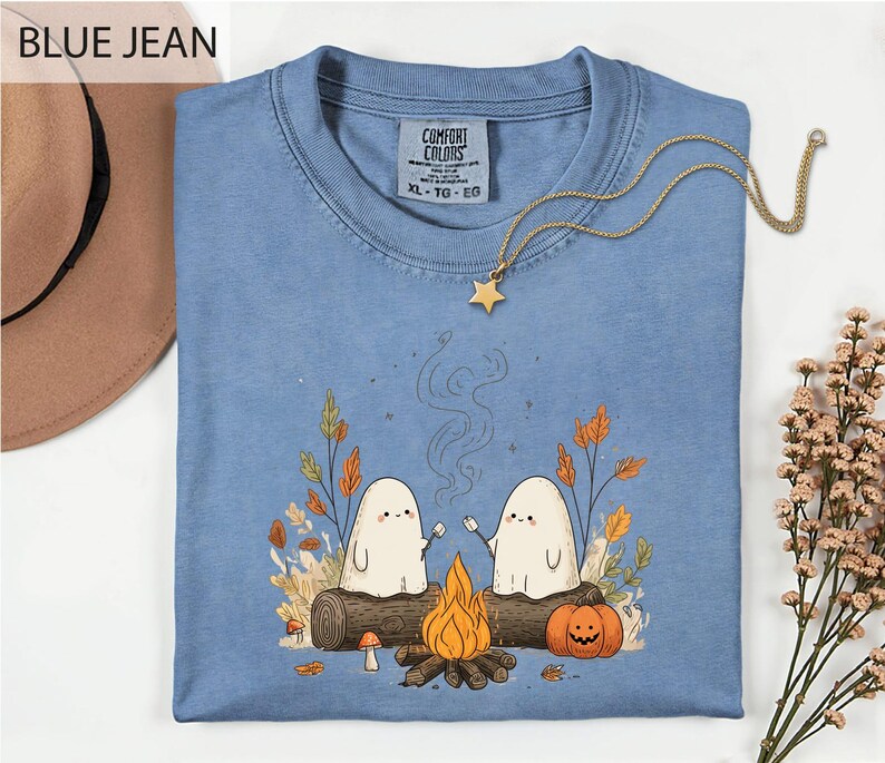 a blue t-shirt with a graphic design depicting two ghostly figures sitting by a campfire, surrounded by autumn foliage and a pumpkin.