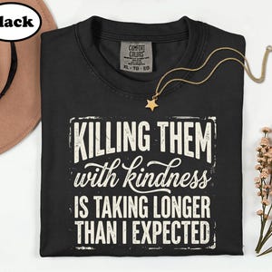 May include: A black t-shirt featuring the white distressed text "KILLING THEM with kindness IS TAKING LONGER THAN I EXPECTED" centered on the chest. The shirt has a Comfort Colors brand label at the inner neckline indicating size XL.