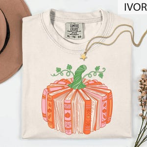 A white t-shirt with a graphic design of a pumpkin made up of books, accompanied by a necklace and a hat, all placed on a white background.