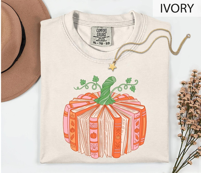 A white t-shirt with a graphic design of a pumpkin made up of books, accompanied by a necklace and a hat, all placed on a white background.