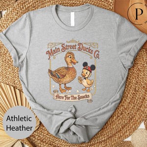 A gray t-shirt with a cartoon image of a duck and a duckling, along with the text "Main Street Ducks Co. Here For The Snacks" printed on it.