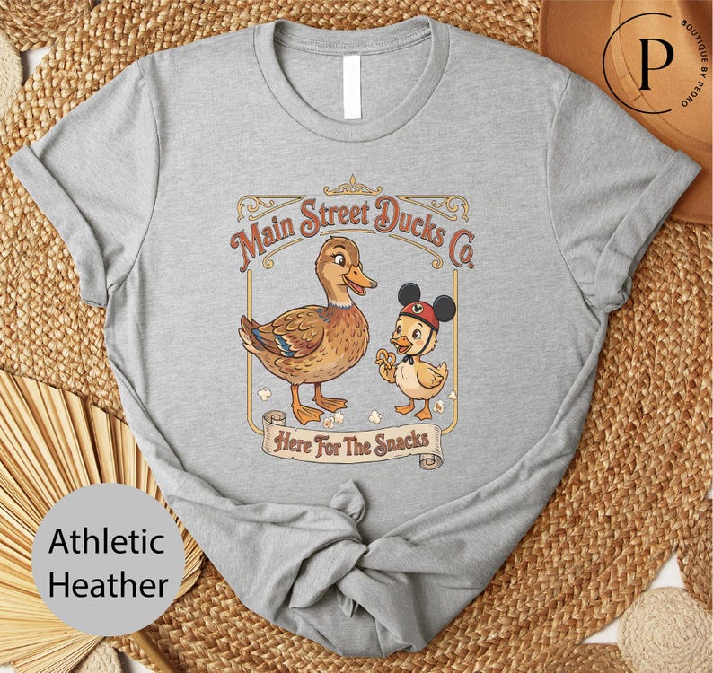 A gray t-shirt with a cartoon image of a duck and a duckling, along with the text "Main Street Ducks Co. Here For The Snacks" printed on it.