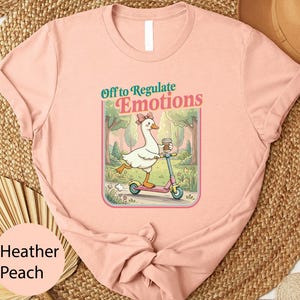 May include: Heather peach t-shirt featuring a cartoon goose riding a pink scooter and holding a coffee cup. The graphic includes the text "Off to Regulate Emotions" in a whimsical font. The shirt is made of a soft material.