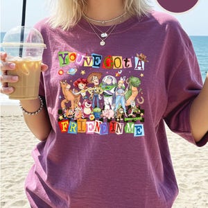 A woman wearing a purple t-shirt with a colorful graphic of characters from Toy Story, holding a drink and standing on a beach.
