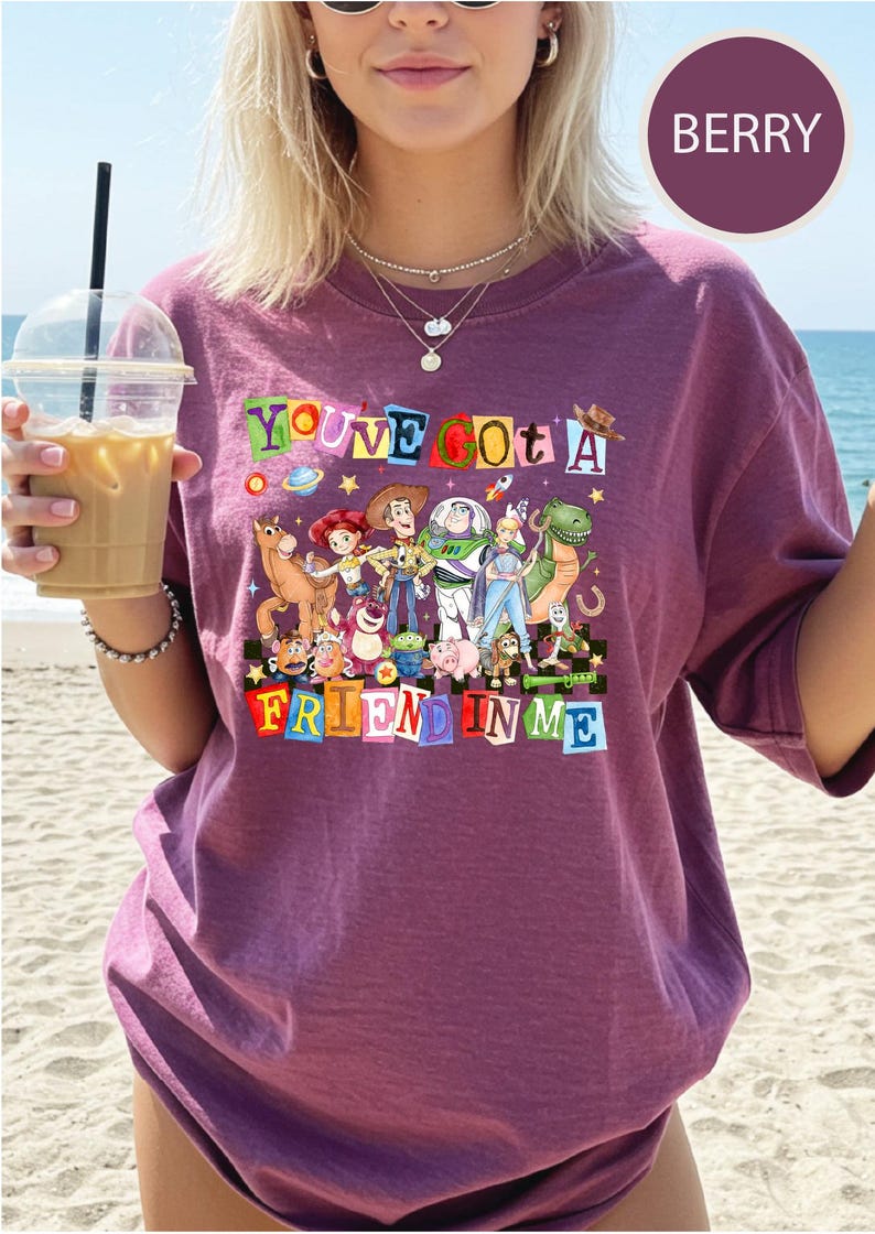 A woman wearing a purple t-shirt with a colorful graphic of characters from Toy Story, holding a drink and standing on a beach.