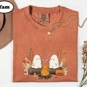 a t-shirt with a Halloween-themed graphic design, including two ghostly figures, a campfire, pumpkins, and a star. The t-shirt is displayed on a white background, accompanied by a hat and some flowers.