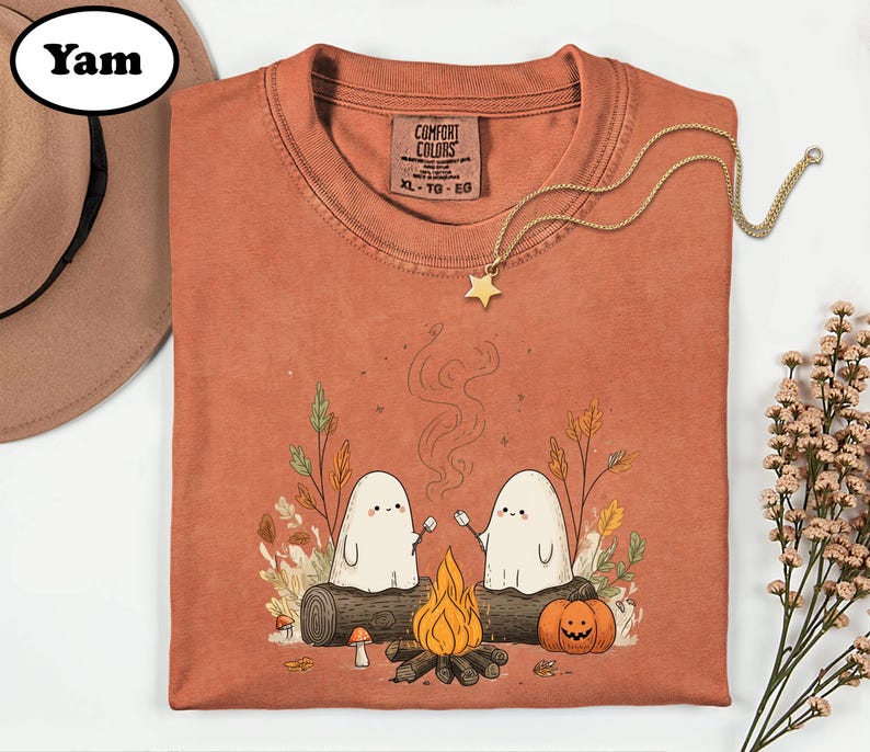 a t-shirt with a Halloween-themed graphic design, including two ghostly figures, a campfire, pumpkins, and a star. The t-shirt is displayed on a white background, accompanied by a hat and some flowers.