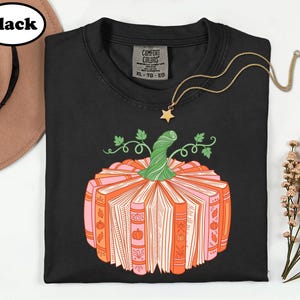 A black t-shirt with a graphic design of a pumpkin made up of books, along with a necklace and a hat.