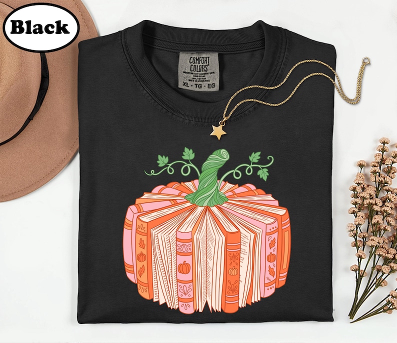 A black t-shirt with a graphic design of a pumpkin made up of books, along with a necklace and a hat.