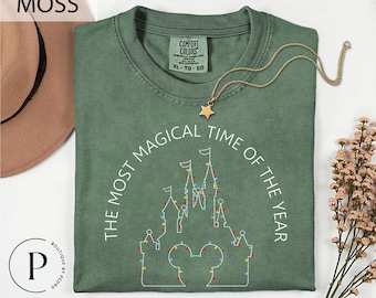 May include: Moss green t-shirt with the text "THE MOST MAGICAL TIME OF THE YEAR" and a castle design with Mickey Mouse ears. The shirt has a crew neck and short sleeves. A gold star pendant necklace is draped across the shirt.