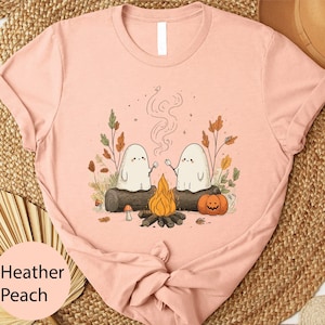 A pink t-shirt with a whimsical graphic design depicting two ghostly figures sitting by a campfire, surrounded by autumn foliage and pumpkins.